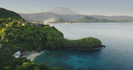 Aerial view secluded Bali Blue Lagoon beach with lush green hills, traditional coastal villages and tranquil turquoise water framed by Mount Agung. Tropical island landscape. Drone flight footageの写真素材