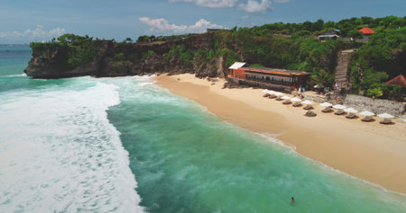 Turquoise waves crashing on Dreamland Beach, Bali, with beach chairs and umbrellas dotting the sandy shore, showcasing the beauty of Indonesia from an aerial perspectiveの写真素材