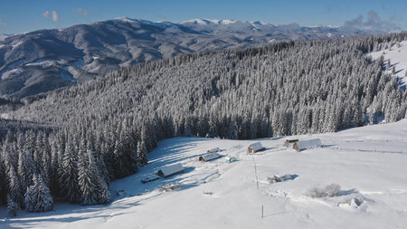 Breathtaking aerial drone revealing serene winter beauty snow covered mountain peaks, dense white blanketed forest, cozy houses nestled among frosty trees. Winter wild nature travel backgroundの写真素材