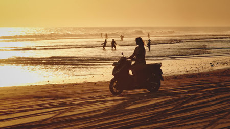 Silhouette of beautiful woman riding scooter along sandy beach, golden warm sunset over ocean waves in Bali, local fishermen fishing in background. Remote wild nature paradise, exotic summer travelの写真素材