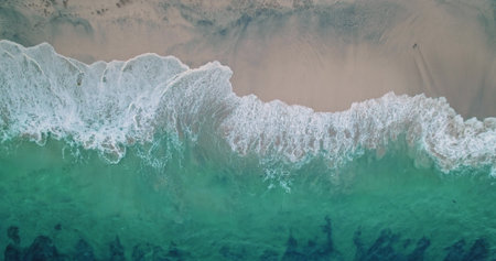 Aerial drone captures the mesmerizing turquoise ocean waves crashing gently on the sandy shore of...