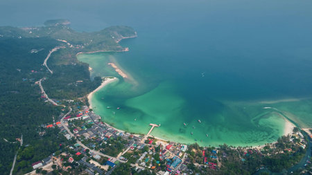 Aerial view capturing a coastal road winding through lush greenery, leading to a vibrant town beside...