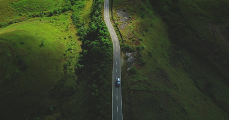 Aerial drone capturing a car driving along a winding road amidst the lush green hills of Sumba...