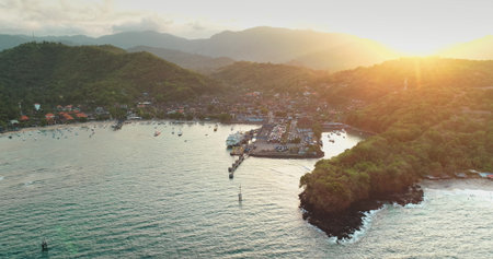 Golden hour sunlight illuminates the picturesque Padang Bai harbor in Bali, Indonesia, showcasing the vibrant convergence of land and sea with boats gently swaying in the tranquil watersの写真素材