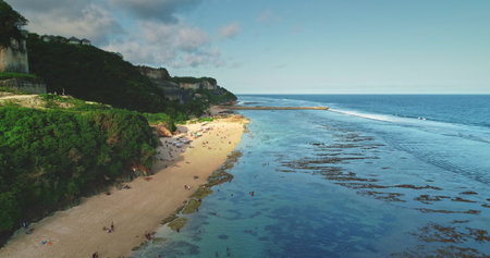 Crystal-clear turquoise water lapping against a sandy shore at a picturesque Bali beach, framed by lush cliffs and bustling tourists enjoying the tropical paradiseの写真素材