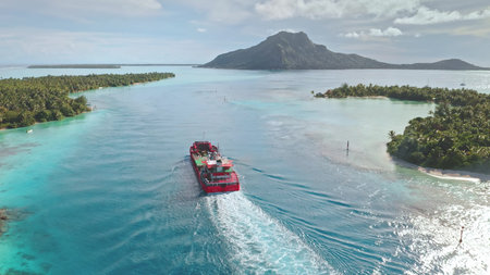 Red cargo ship sailing turquoise water lagoon, passing near tropical island with lush vegetation...