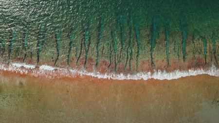An aerial perspective showcases a calm shoreline where gentle waves meet the sandy beach. The interplay of water and land creates a tranquil scene during sunset hours.の写真素材