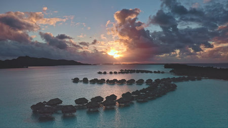 Dramatic colorful sunset sky over Bora Bora with overwater bungalows silhouette, turquoise lagoon water, tropical island in background. Remote wild nature paradise, romantic honeymoon. Aerial viewの写真素材