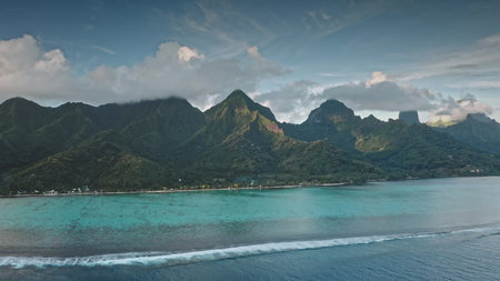 Aerial panorama flight over Moorea tropical island, French Polynesia. Green mountains range towering over turquoise coral reef lagoon ocean under blue clouds sky. Remote wild nature travel. Drone viewの写真素材