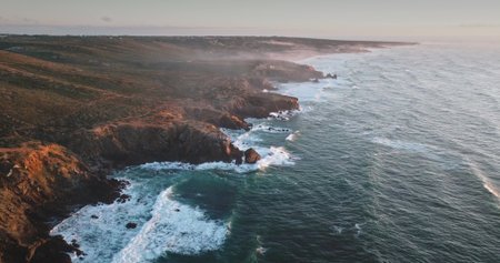 Portugal, Lisbon: Dramatic rugged cliffs and verdant hills lining the Atlantic Ocean coastline, with golden hour sunlight illuminating the landscape, waves crash against the shore. Aerial drone flightの写真素材