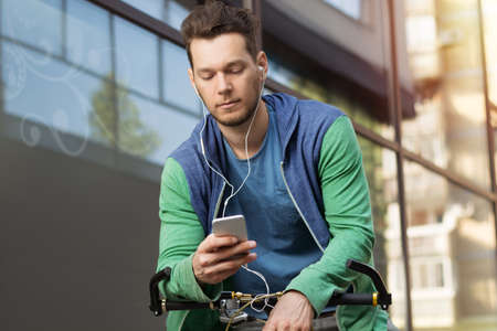 Young man in city checking his smartphone while sitting on a bicycleの写真素材