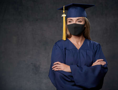 Young woman graduate in black mask with hands crossed celebrating her university degree wearing blue cap and gown looking to the sideの写真素材