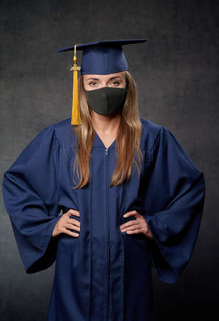 Young woman graduate both hands on the side celebrating her university degree wearing blue cap and gown also wearing black maskの写真素材