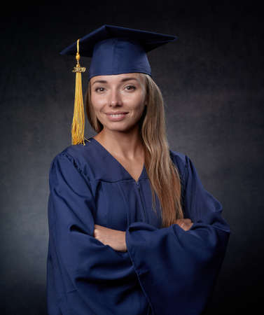 Smiling young woman arms crossed wearing blue cap and gownの写真素材