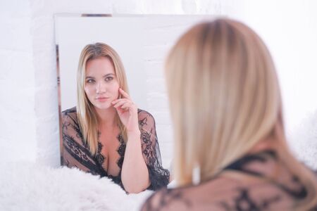 charming plus size caucasian blonde woman dressed in beautiful lace black lingerie and looking at herself in the mirror at home aloneの写真素材