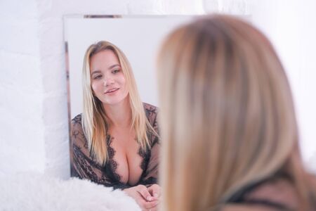 charming plus size caucasian blonde woman dressed in beautiful lace black lingerie and looking at herself in the mirror at home aloneの写真素材