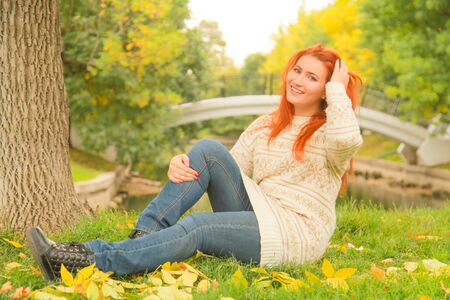 redhaired woman in a warm white autumn sweater sitting on the ground near a tree in a city Parkの写真素材