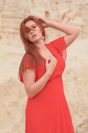 Young beautiful Caucasian woman in long red dress posing in desert landscape with sand.の写真素材