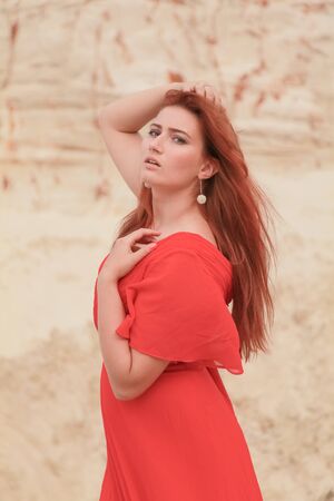 Young beautiful Caucasian woman in long red dress posing in desert landscape with sand.の写真素材