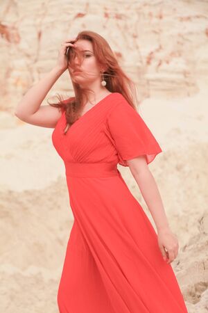 Young beautiful Caucasian woman in long red dress posing in desert landscape with sand.の写真素材