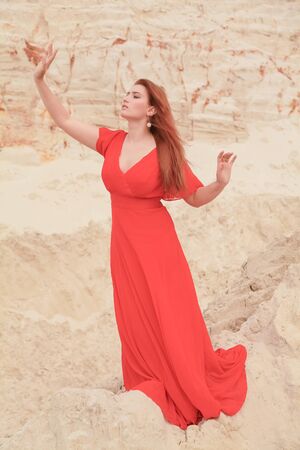 Young beautiful Caucasian woman in long red dress posing in desert landscape with sand.の写真素材