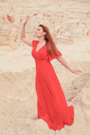 Young beautiful Caucasian woman in long red dress posing in desert landscape with sand.の写真素材