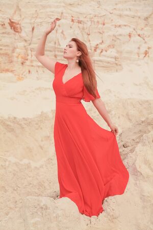 Young beautiful Caucasian woman in long red dress posing in desert landscape with sand.の写真素材