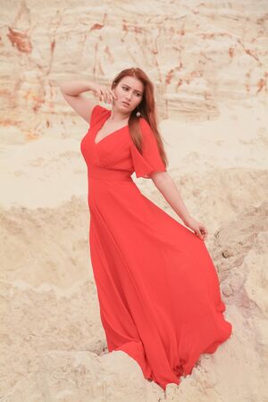 Young beautiful Caucasian woman in long red dress posing in desert landscape with sand.の写真素材