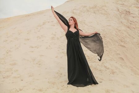 Young beautiful Caucasian woman in black chiffon dress posing in desert landscape with sand.の写真素材