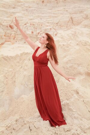 Young beautiful plus size Caucasian woman in red long dress posing in desert landscape with sand.の写真素材