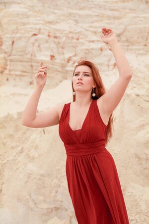 Young beautiful plus size Caucasian woman in red long dress posing in desert landscape with sand.の写真素材