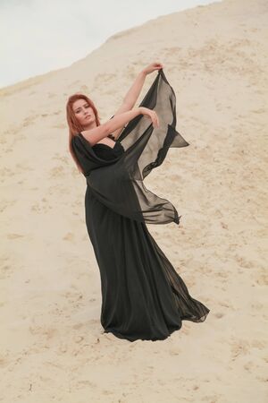 Young beautiful Caucasian woman in black chiffon dress posing in desert landscape with sand.の写真素材