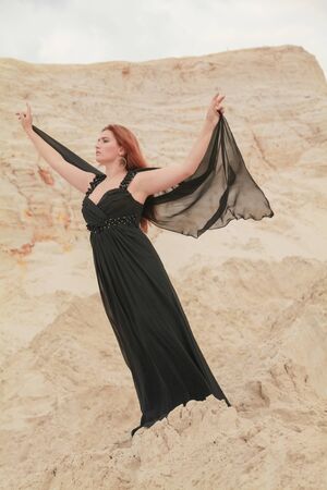 Young beautiful Caucasian woman in black chiffon dress posing in desert landscape with sand.の写真素材