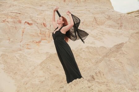 Young beautiful Caucasian woman in black chiffon dress posing in desert landscape with sand.の写真素材