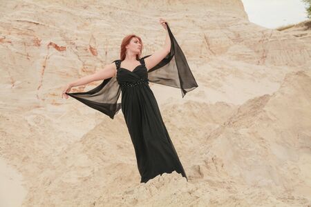 Young beautiful Caucasian woman in black chiffon dress posing in desert landscape with sand.の写真素材