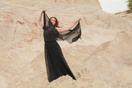 Young beautiful Caucasian woman in black chiffon dress posing in desert landscape with sand.の写真素材