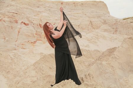 Young beautiful Caucasian woman in black chiffon dress posing in desert landscape with sand.の写真素材