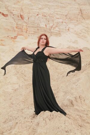 Young beautiful Caucasian woman in black chiffon dress posing in desert landscape with sand.の写真素材