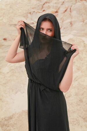 Young beautiful Caucasian woman in black chiffon dress posing in desert landscape with sand.の写真素材