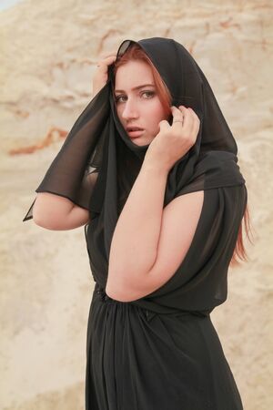 Young beautiful Caucasian woman in black chiffon dress posing in desert landscape with sand.の写真素材