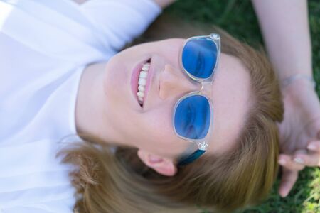 pretty caucasian girl in blue sunglasses lying on the green grass and laugh happyの写真素材
