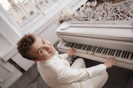 young man teenager in a white business suit with a large white piano in flowers. caucasian male play...