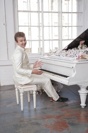 young man teenager in a white business suit with a large white piano in flowers. caucasian male play music at holiday alone.の写真素材