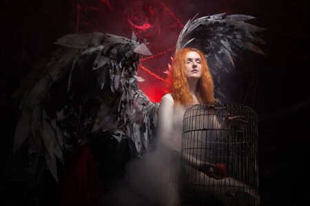 an evil tempting woman with large demon wings holds an Apple in a large cage and beckons to sin. Halloween photo plus size girl with red hair on a huge Gothic throne.の写真素材