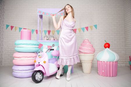cute happy adult girl posing in fashionable dress among huge fabulous sweets and cakesの写真素材