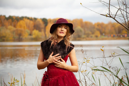 Young woman in stylish hat and long red evening dress with fur posing near the river and walking in the autumn garden background aloneの写真素材