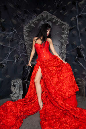 beautiful vampire woman in red long dress near big black throne in the studioの写真素材