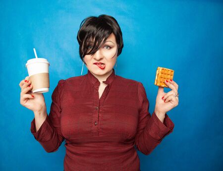 joyful plus size woman with short hair in business clothes resting and drinking coffee with a straw with Viennese waffle as snack on a blue studio background.の写真素材