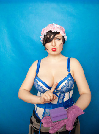 adorable happy plus size short hair woman in blue white lingerie with a rubber glove and a sponge for washing dishes on a solid background in Studioの写真素材