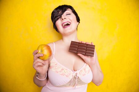 an attractive plus size girl weighs her options and decide to eat chocolate or an apple on bright yellow studio backgroundの写真素材